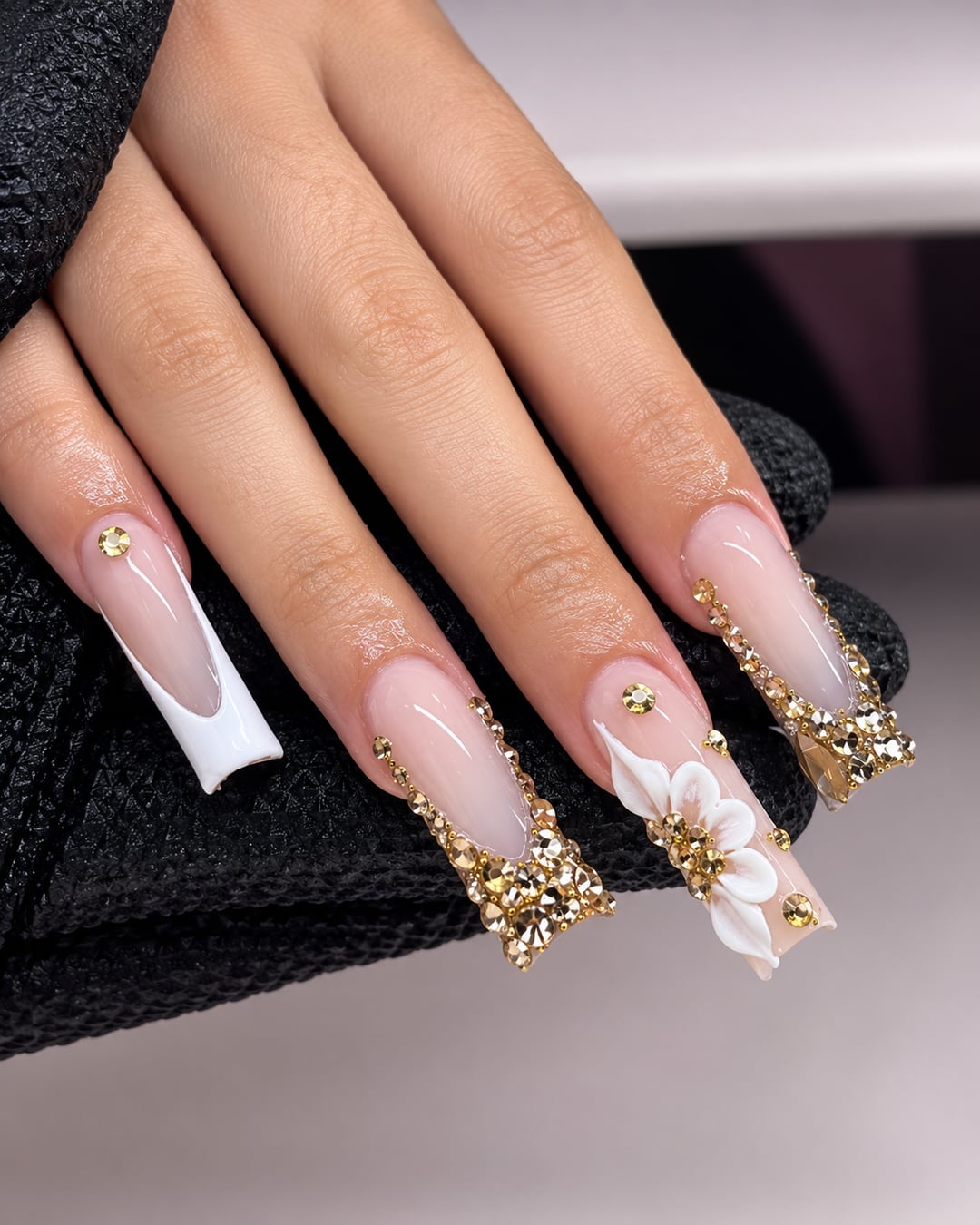Manucure nude et blanche avec strass dorés et fleur 3D, mise en valeur par des guirlandes lumineuses